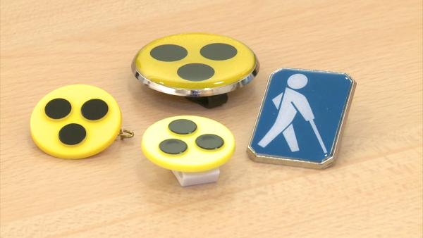 Gelber Blindenanstecker mit schwarzen Punkten, weißes Stockmännchen auf blauem Grund