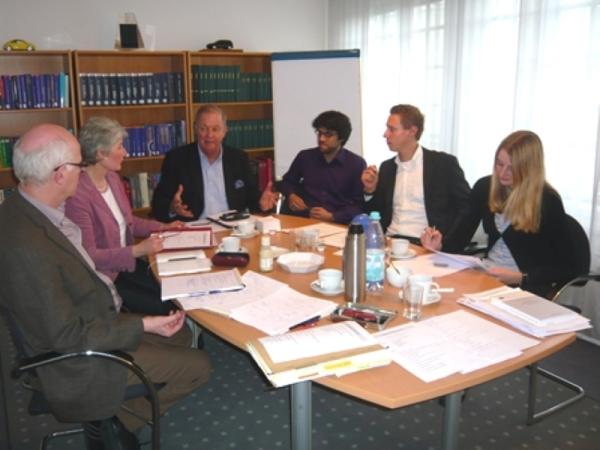 An einem Tisch, mit vielen Papieren und Getränken, sitzt eine Gruppe und hört einem Referenten zu.