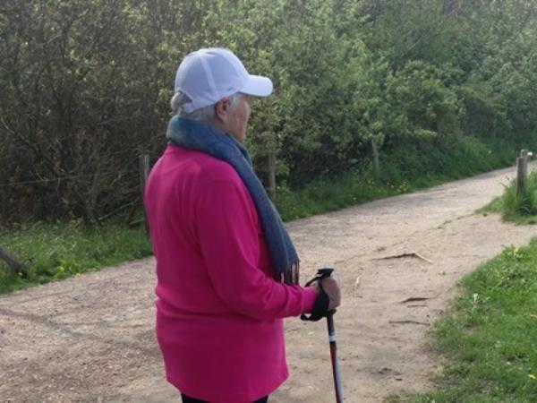 Eine ältere Frau steht auf einem Spazierweg. In den Händen hat sie Wanderstöcke und auf dem Kopf trägt sie eine Schirmmütze.