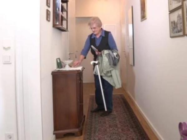 Eine älter Frau steht im Flur einer Wohnung und legt etwas auf ein schmales Schränkchen. Über dem Arm hängt ihre Jacke und am Handgelenk hängt ein weißer Gehstock.  