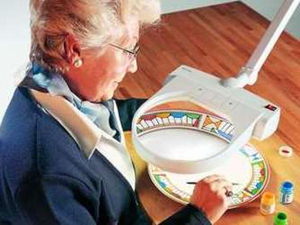 Eine Frau sitzt an einem Tisch und schaut durch eine Lupe mit einer hellen Beleuchtung auf einen Teller, den sie mit einem feinen Pinsel bemalt.  