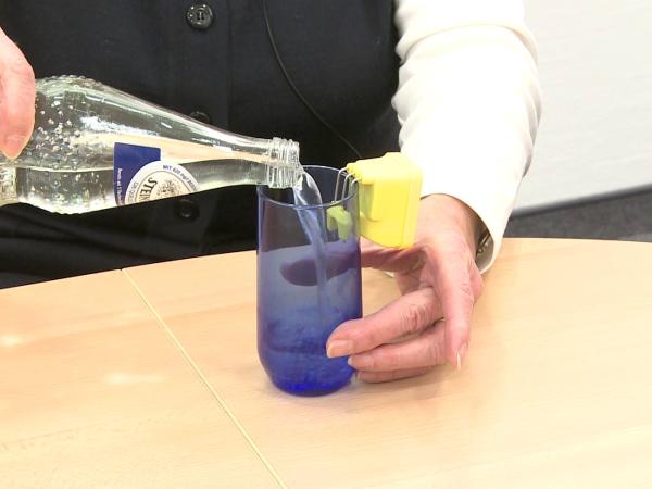 In ein blaues Trinkglas wird Mineralwasser eingegossen. Am Glasrand hängt ein gelber Füllstandsanzeiger.