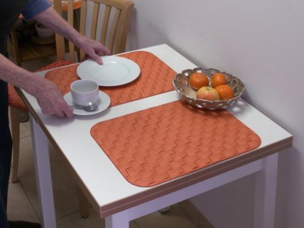 Kontraste Esstisch Auf einem weißen Tisch liegen zwei orange Platzsets. Auf dem einem Platzset wird ein weißer Teller und eine weiße Tasse mit Untertasse gestellt. Ein Metallkorb mit 4 Mandarinen steht in der Mitte des Tisches.