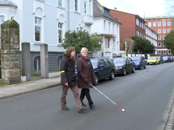 Eine ältere Frau mit Blindenlangstock überquert in Begleitung einer anderen Frau eine Straße.