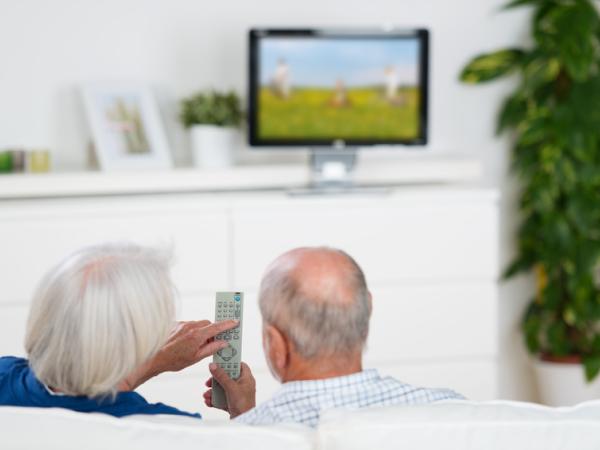 Zwei ältere Menschen sitzen vor einem Fernseher und schauen auf die Fernbedienung des Geräts.