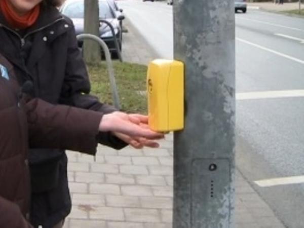 Zwei Personen stehen vor einem Ampelmast mit einem gelben Signalgeber und betätigen die Anforderungstaste.