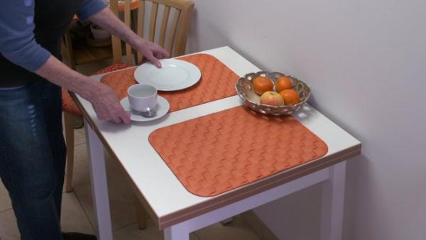 Kontraste Esstisch Auf einem weißen Tisch liegen zwei orange Platzsets. Auf dem einem Platzset wird ein weißer Teller und eine weiße Tasse mit Untertasse gestellt. Ein Metallkorb mit 4 Mandarinen steht in der Mitte des Tisches.